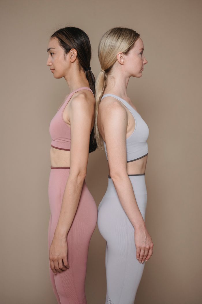 Two women in colorful activewear standing back to back in a neutral studio setting.